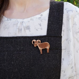 Könnte beinhalten: Ein hölzerner Widder-Anstecker mit detailliertem Design, braunem Körper und gebogenen Hörnern. Die Brosche ist an einem dunkelgrauen Wollkleidungsstück befestigt, vor einer weißen Bluse mit Schafmuster.