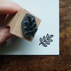 Op de afbeelding: Een houten stempel met een bladeren ontwerp wordt boven een wit oppervlak gehouden, naast een gestempelde afbeelding van hetzelfde blad. De stempel heeft een zwart bladeren patroon en een houten handvat. De gestempelde afbeelding is zwart.