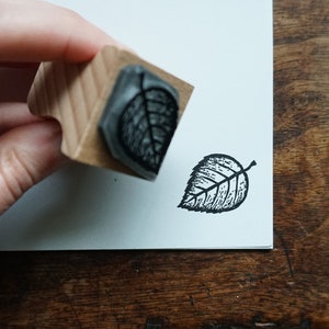 May include: A wooden leaf stamp creating a black ink impression on white paper. The stamp is held, showing the leaf design. The stamped leaf is approximately 3.8 cm in diameter, with visible veins and a detailed pattern.