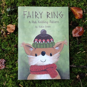 Op de afbeelding: Een boekomslag getiteld "FAIRY RING" met de ondertitel "A Hat Knitting Pattern" door Katie Green. De omslag toont een cartoonvos met een hoed met paddenstoelontwerpen en een rode sjaal. De achtergrond is groen.