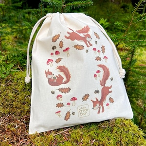May include: Cream-colored drawstring bag featuring a whimsical design of squirrels, mushrooms, and leaves. The squirrels are depicted in various poses, surrounded by red-capped mushrooms and autumn leaves. The bag has a natural, outdoorsy theme.