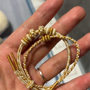 Könnte beinhalten: Ein geflochtenes Armband in Weiß und Gold mit einem Knotendesign. Das Armband besteht aus zwei Schnüren, einer weißen und einer goldenen, die miteinander geflochten sind.