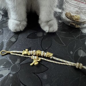 Könnte beinhalten: Ein weißes und goldenes Perlenarmband mit einem goldenen Anhänger in Form eines kleinen Tieres. Das Armband liegt auf einer schwarz-weißen gemusterten Oberfläche.