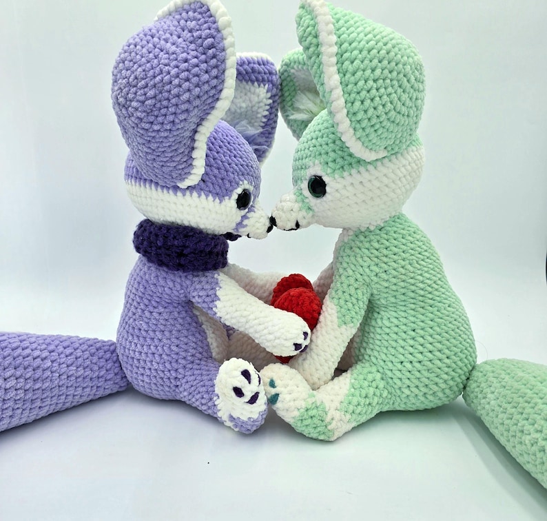 Pu&ograve; includere: Due peluche a forma di volpe all'uncinetto, uno lavanda e l'altro verde menta, con dettagli bianchi. La volpe lavanda indossa una sciarpa viola scuro. Tengono un piccolo cuore rosso. I peluche hanno grandi orecchie e occhi neri.