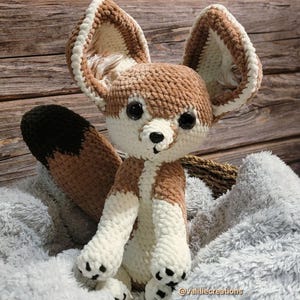 May include: A handmade crochet Fennec fox plush toy. The fox has large, pointed ears with white and brown accents, a brown and white body, and a black-tipped tail. It has black eyes and a black nose. The plush is sitting on a grey, fluffy blanket.