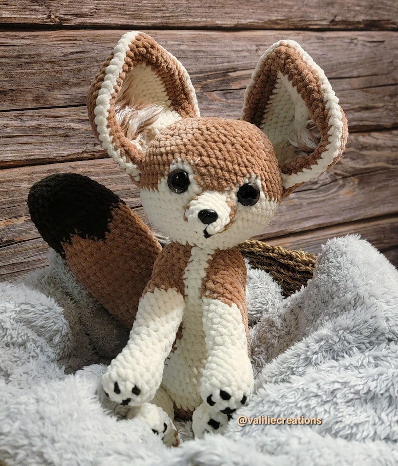 Pu&ograve; includere: Un peluche fatto a mano a uncinetto a forma di volpe Fennec. La volpe presenta grandi orecchie a punta con accenti bianchi e marroni, un corpo marrone e bianco e una coda con la punta nera. Ha occhi neri e un naso nero. Il peluche &egrave; seduto su una coperta grigia e soffice.