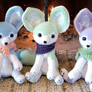 Pu&ograve; includere: Tre peluche a forma di volpe fatti a mano all'uncinetto, blu, verdi e viola. Ogni volpe ha grandi orecchie, una coda soffice e una sciarpa. I peluche sono su una superficie tessuta davanti a un camino.