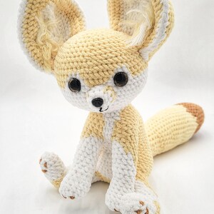 Pu&ograve; includere: Un peluche di volpe Fennec all'uncinetto in crema, bianco e beige. L'animale di peluche fatto a mano ha grandi orecchie, una lunga coda e occhi scuri. La volpe &egrave; seduta in posizione eretta, mostrando i suoi dettagli.