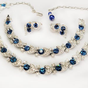May include: A vintage jewelry set featuring a necklace, bracelet, and earrings. The set is silver-toned with clear rhinestones and deep blue, round gemstones. The necklace and bracelet have a leaf and floral design.