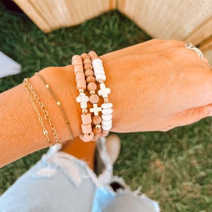 Könnte beinhalten: Ein Set aus vier Armbändern mit Holzperlen und kleinen goldenen Kreuzanhängern. Die Armbänder sind auf dem Handgelenk einer Person übereinander gelegt.