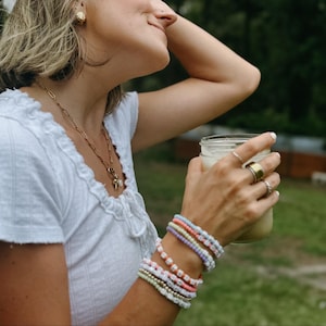 May include: A collection of colorful beaded bracelets in pastel shades, including pink, blue, green, and white. The bracelets are stacked on a wrist, with a gold ring and a clear glass jar with a white liquid in the background.