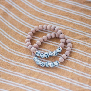 May include: Two bracelets, one with light brown wooden beads and the other with white beads with a blue floral pattern.