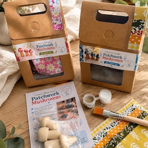 May include: Two brown cardboard boxes labelled Create Your Own Patchwork Mushrooms DIY Craft Kit. Each box has a cutout handle and a window. Included are a printed instruction sheet, four wooden mushroom shapes, two paint pots, and a paintbrush.