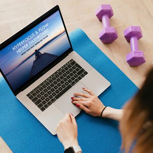 Puede incluir: Un portátil que muestra "HYPERFOCUS & FLOW HELPER DASHBOARD" está sobre una esterilla de yoga azul. Dos pesas moradas y una botella de agua están cerca. La pantalla muestra a una persona en un muelle al atardecer. Manos en el portátil.