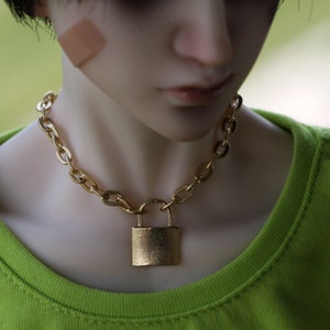 May include: A gold chain necklace with a padlock pendant. The necklace is worn by a person with short black hair.