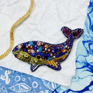 May include: A blue and gold whale brooch with colorful beads and sequins. The brooch is on a white background with blue marbled paper.