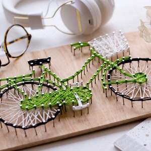 May include: A green and white string art bicycle with a basket on the handlebars. The bicycle is mounted on a wooden board.