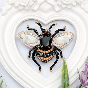 Peut inclure: Une broche en forme d'abeille noire et dorée avec des ailes blanches, faite de perles et de strass. La broche est présentée dans un cadre blanc en forme de cœur.