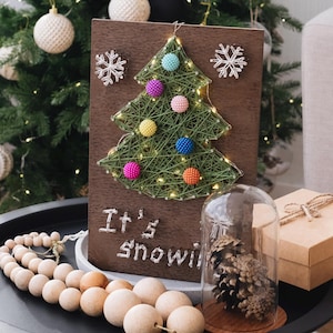 May include: A wooden sign with a string art Christmas tree design. The tree is decorated with colorful beads and lights. The sign also says "It's Snowing".