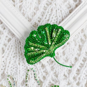 Op de afbeelding: Een groene ginkgo-blad broche gemaakt van kralen en strass. De broche ligt op een witte gebreide stoffen achtergrond.