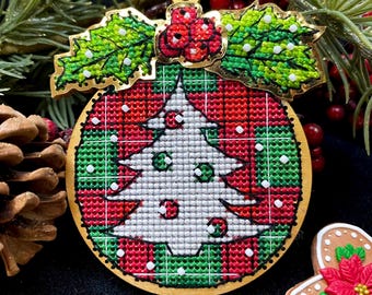 May include: A round wooden Christmas bauble with a red and green plaid pattern and a white embroidered Christmas tree. The bauble is decorated with green holly leaves and red berries.