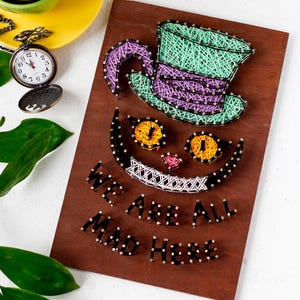 Puede incluir: Arte de hilo del Gato de Cheshire de Alicia en el País de las Maravillas. La cara del gato está hecha de hilo negro, blanco y amarillo. El gato lleva un sombrero verde y morado. El texto "We are all mad here" está escrito debajo de la cara del gato.