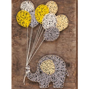 May include: A gray string art elephant holding a bunch of yellow and white string art balloons. The elephant and balloons are on a brown wooden background.