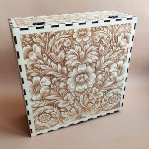 Könnte beinhalten: Eine Holzkiste mit einem detaillierten Blumenmuster, das in die Oberfläche geschnitzt ist. Die Kiste ist hellbraun mit einem cremefarbenen Rahmen und Deckel. Das florale Design umfasst Blumen und Blätter und schafft einen dekorativen Aufbewahrungsbehälter.