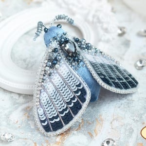 May include: A blue velvet moth brooch with silver and black sequins, rhinestones, and pearls. The moth has a large, iridescent black gemstone in the center of its body.