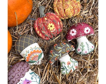 May include: A collection of fall-themed fabric ornaments, including pumpkins, acorns, and mushrooms, with intricate embroidery and beadwork. The ornaments are in shades of red, orange, yellow, and brown, and are arranged on a bed of straw.