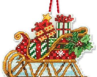 May include: A red and green Christmas ornament featuring a sleigh filled with presents and a sprig of holly.