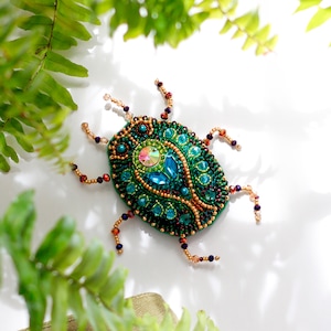 May include: A green and blue jeweled beetle brooch with a gold and brown beaded outline. The brooch is on a white background.