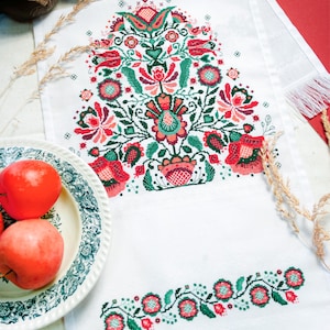 May include: White embroidered table runner with a vibrant floral design in red, green, and coral. The runner features a large central floral motif and a smaller border pattern. The design is reminiscent of traditional folk art.