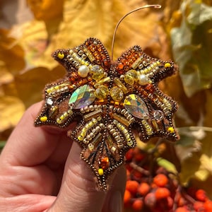 Könnte beinhalten: Handgefertigte Brosche in Blattform, herbstlich gestaltet. Sie ist mit braunen, goldenen und gelben Perlen verziert, ergänzt durch schillernde Kristalle. Ein feiner Drahtstiel verstärkt das realistische Design. Ein schönes Accessoire.