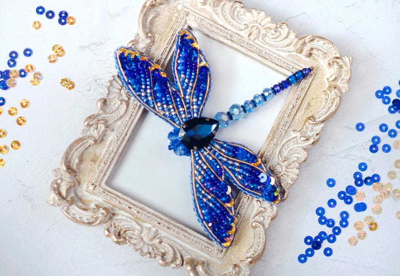 Puede incluir: Un broche de lib&eacute;lula azul y dorado con una gema negra en el centro, exhibido en un marco blanco adornado.
