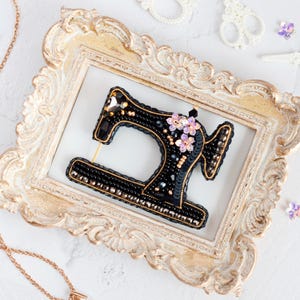 May include: A black beaded sewing machine brooch with gold accents and floral embellishments. The brooch is set inside a vintage gold frame. Additional sewing-themed accessories are scattered around the frame.