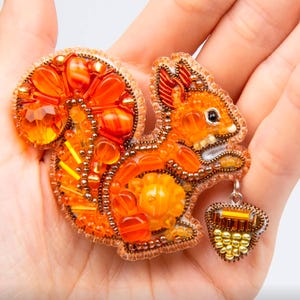 May include: A detailed, orange beaded squirrel brooch with a matching acorn charm. The brooch features various shades of orange beads and gems, with a detailed design and a silver-colored charm. The squirrel is holding an acorn.