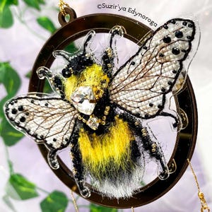 May include: A black and yellow bumblebee with white fur, beaded wings, and a crystal embellishment is displayed in a round, clear, acrylic frame with a gold-colored metal rim. The frame is suspended from a gold chain.