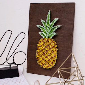 Op de afbeelding: Een string art ananas met een geel lichaam en groene bladeren op een bruine houten achtergrond.