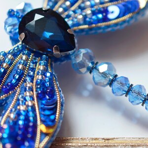 Puede incluir: Un broche de lib&eacute;lula azul con una gran piedra preciosa azul oscuro en el centro. Las alas est&aacute;n hechas de cuentas y lentejuelas azules y plateadas. El broche est&aacute; unido a una cadena de cuentas de vidrio azules.