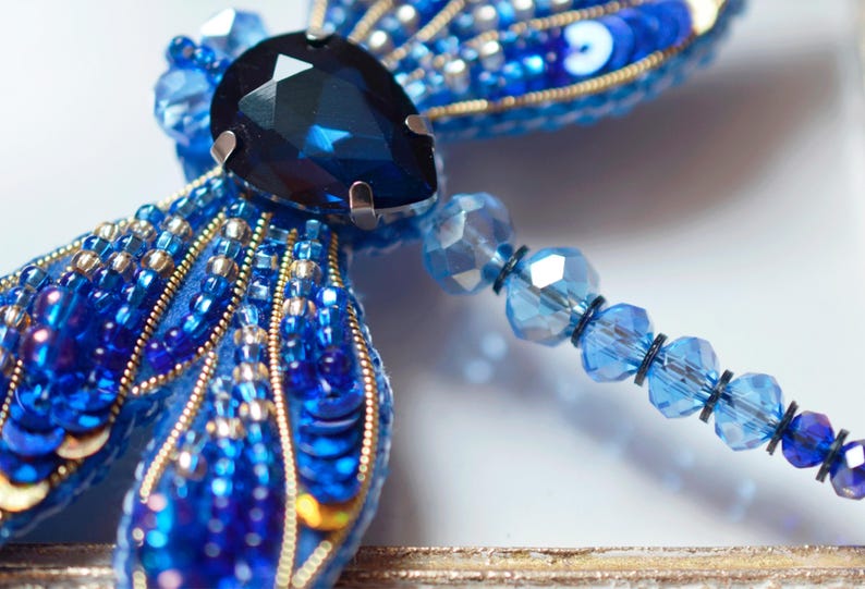 Puede incluir: Un broche de lib&eacute;lula azul con una gran piedra preciosa azul oscuro en el centro. Las alas est&aacute;n hechas de cuentas y lentejuelas azules y plateadas. El broche est&aacute; unido a una cadena de cuentas de vidrio azules.