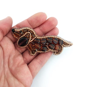 Op de afbeelding: Handgemaakte teckelbroche, met een kralenontwerp in bruin- en amberkleuren. De hondenpin is gemaakt met kleine, ronde kralen en grotere, ovaalvormige kralen, waardoor een getextureerd en gedetailleerd uiterlijk ontstaat. De broche is een uniek accessoire.