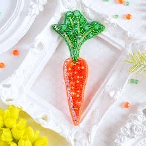 May include: A beaded carrot brooch with a green leafy top and an orange body. The brooch is displayed on a white surface with a white frame.