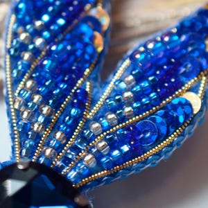 Puede incluir: Primer plano de un broche azul y dorado con cuentas, con una gran piedra preciosa azul oscuro en el centro. El broche est&aacute; hecho con peque&ntilde;as cuentas y lentejuelas iridiscentes.
