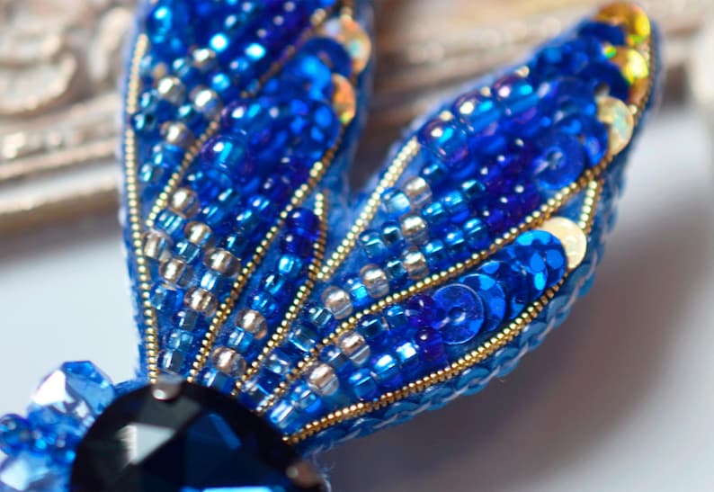 Puede incluir: Primer plano de un broche azul y dorado con cuentas, con una gran piedra preciosa azul oscuro en el centro. El broche est&aacute; hecho con peque&ntilde;as cuentas y lentejuelas iridiscentes.