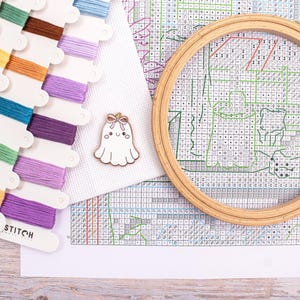 May include: A cross-stitch kit with colorful thread spools, a wooden embroidery hoop, and a white fabric with a cross-stitch pattern. A small, white ghost-shaped pin with a pink bow is also present. The kit is for a craft project.
