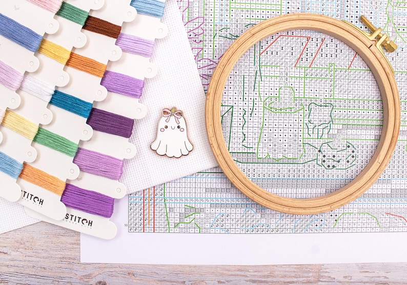 May include: A cross-stitch kit with colorful thread spools, a wooden embroidery hoop, and a white fabric with a cross-stitch pattern. A small, white ghost-shaped pin with a pink bow is also present. The kit is for a craft project.