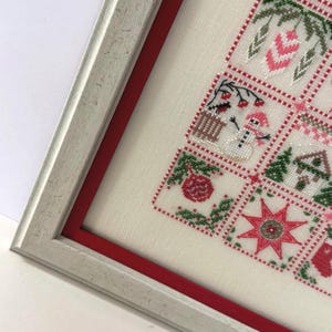 May include: Framed cross-stitch artwork featuring a Christmas tree design. The artwork is in a white frame with a red inner border. The cross-stitch includes festive elements like ornaments, a snowman, and a star, in red, green, and white.