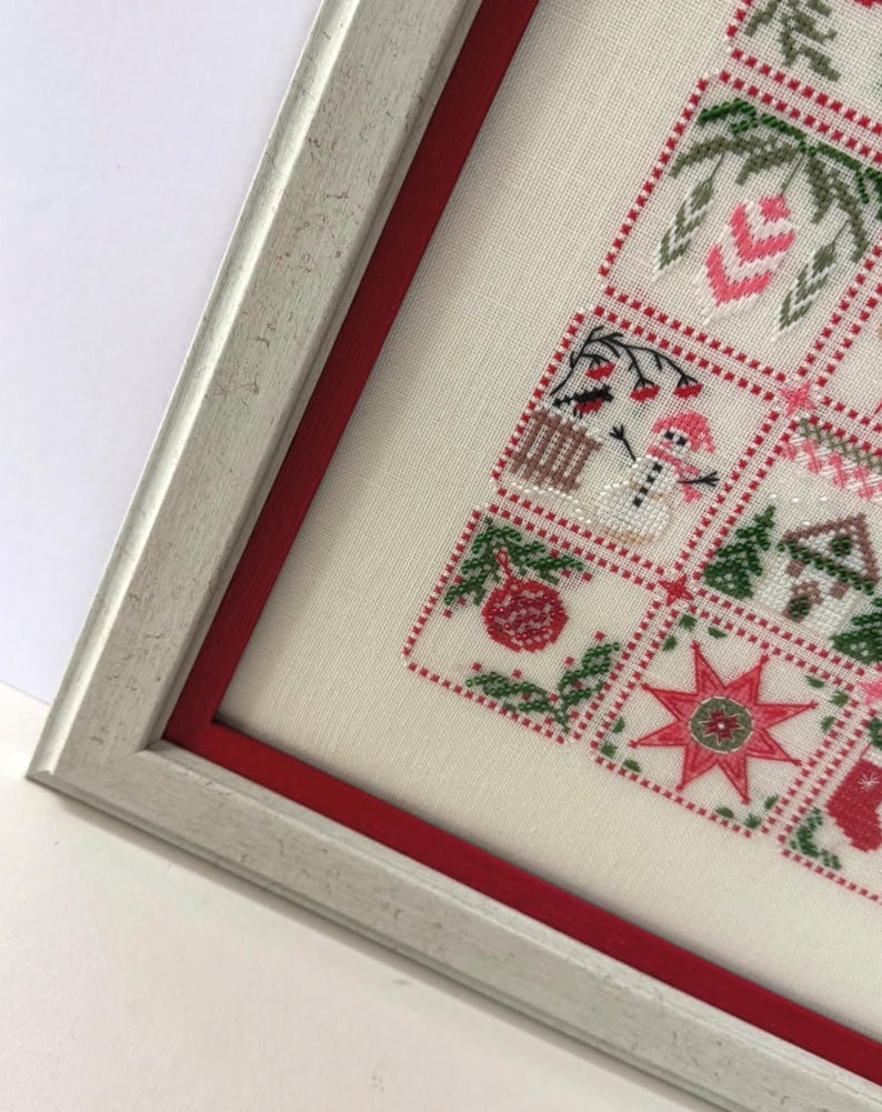 May include: Framed cross-stitch artwork featuring a Christmas tree design. The artwork is in a white frame with a red inner border. The cross-stitch includes festive elements like ornaments, a snowman, and a star, in red, green, and white.