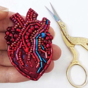 May include: A handmade, beaded heart-shaped brooch in shades of red, with blue and pink accents. The brooch is held in a hand, next to a pair of gold-coloured scissors. The brooch is likely a piece of wearable art.